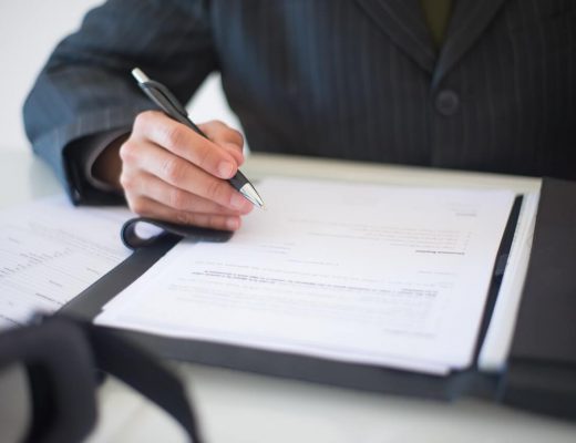 Imagem do braço de um homem segurando uma caneta prestes a assinar um documento em uma mesa. Essa imagem representa um ato de assinatura em cartório de notas.