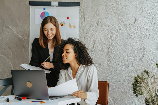 A imagem mostra duas mulheres em frente a um laptop, uma mulher branca de blazer preto em pé e uma mulher negra de roupas brancas sentada. Ambas seguram papéis e apontam para a tela. Essa imagem representa a informatização de cartórios.